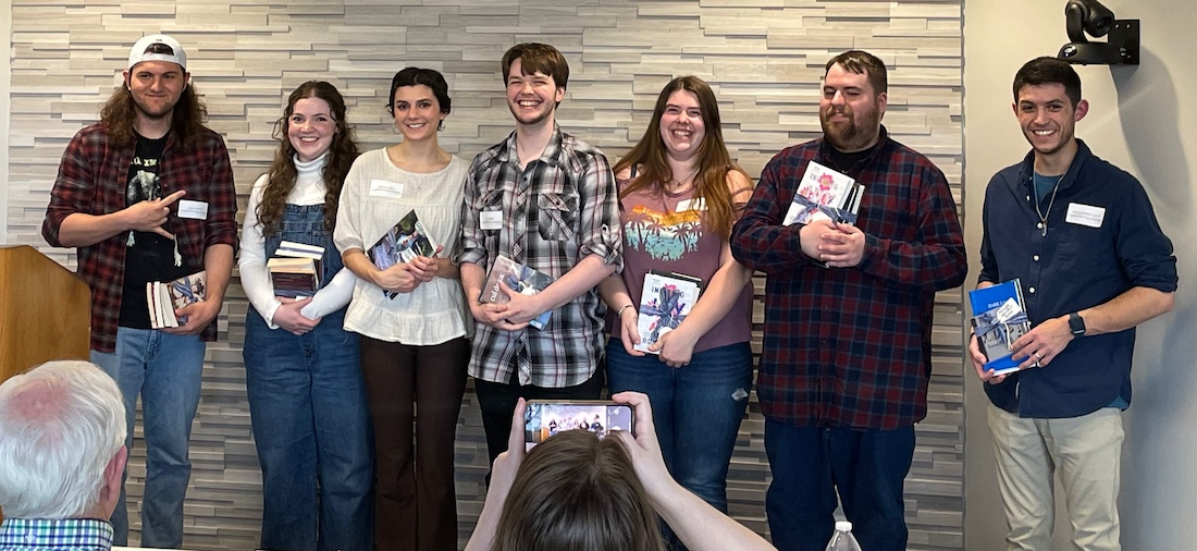 Seven students standing, holding books, and smiling while audience member takes a photo Seven students standing, holding books, and smiling while audience member takes a photo