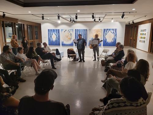 Circle of people sitting in art gallery and listening to one person read and another play guitar